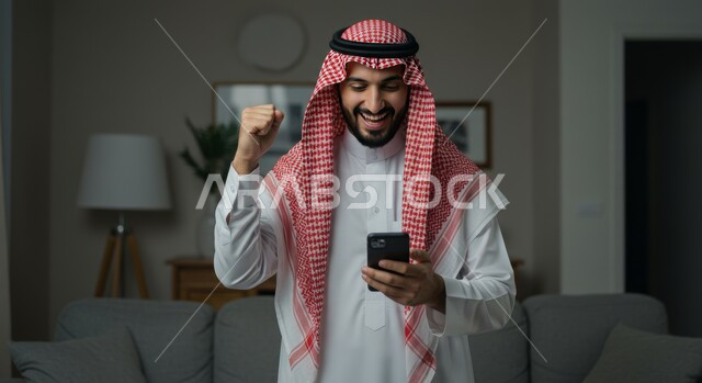 تعابير النصر والفوز، صورة مقربة لرجل عربي خليجي سعودي يرتدي الشماغ والثوب التقليدي يمسك بيده الهاتف المحمول يرفع قبضة يده للاعلى بايماءات الحماس والسرور، رؤية الاخبار المفرحة والسعيدة، تحقيق الاهداف والانجاز