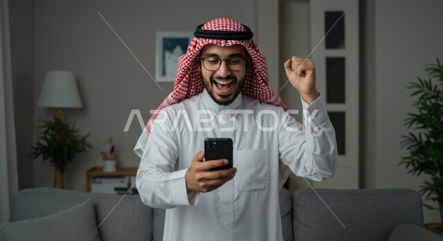 تحقيق الاهداف والانجاز، تعابير النصر والفوز، صورة مقربة لرجل عربي خليجي سعودي يرتدي الشماغ والثوب التقليدي يمسك بيده الهاتف المحمول يرفع قبضة يده للاعلى بايماءات الحماس والسرور، رؤية الاخبار المفرحة والسعيدة