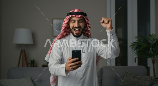 تعابير النصر والفوز، صورة مقربة لرجل عربي خليجي سعودي يرتدي الشماغ والثوب التقليدي يمسك بيده الهاتف المحمول يرفع قبضة يده للاعلى بايماءات الحماس والسرور، رؤية الاخبار المفرحة والسعيدة، تحقيق الاهداف والانجاز