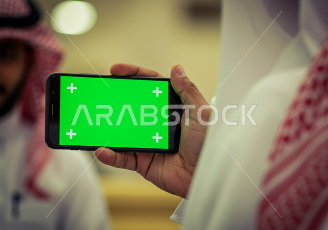 استخدام جهاز تقني حديث متطور، صورة مقربة ليد لرجل عربي خليجي سعودي يرتدي الشماغ والثوب التقليدي يمسك بيده هاتف محمول بشاشة خضراء فارغة، كروما