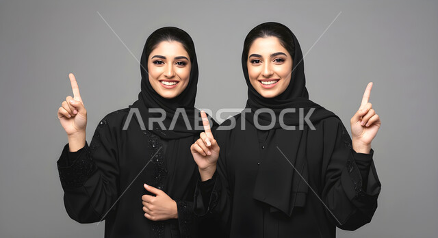 الاشارة بأصبع السبابه نحو الأعلى بإيماءات السرور، الوقوف باستقامة وثقة بالنفس، الاناقة والاهتمام بالمظهر الخارجي، بورتريه مقرب لأختين توءم عربيتين خليجيتين سعوديتين محجبتين مبتسمتين ترتديان العباءة السوداء تنظران الى الكاميرا، خلفية رمادية