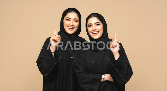 الاناقة والاهتمام بالمظهر الخارجي، الوقوف باستقامة وثقة بالنفس، بورتريه مقرب لأختين توءم عربيتين خليجيتين سعوديتين محجبتين مبتسمتين ترتديان العباءة السوداء تنظران الى الكاميرا، الاشارة بأصبع السبابه نحو الأعلى بإيماءات السرور، خلفية بيج