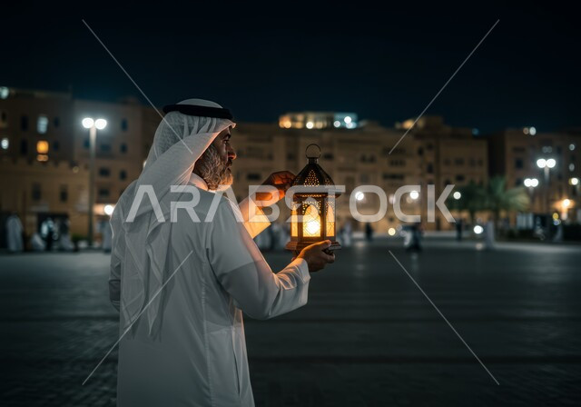 زينة وأنوار الأعياد والمناسبات الدينية الاسلامية، سوق وحي سكني شعبي مضيء في الليل،الاستعداد والتجهز لاستقبال شهر رمضان المبارك، الاحتفال بقدوم شهر الخير، صورة من الخلف لرجل عربي خليجي سعودي يرتدي الغترة والثوب التقليدي يحمل فانوس معدني مزخرف مضيء