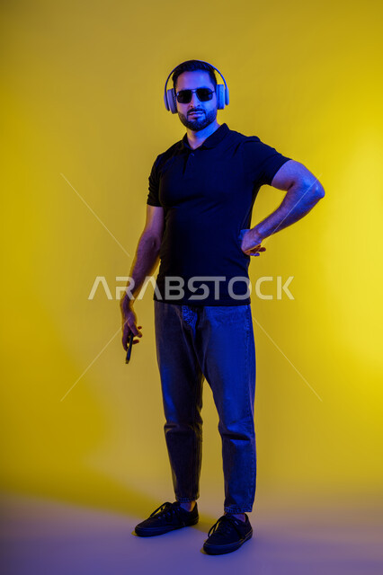 Using headphones, modern advanced technology, making a video call with friends with happy expressions, side portrait of a young Saudi Arabian man in casual wear using a mobile phone and taking a selfie, Emirati man listening to music and songs, yellow background