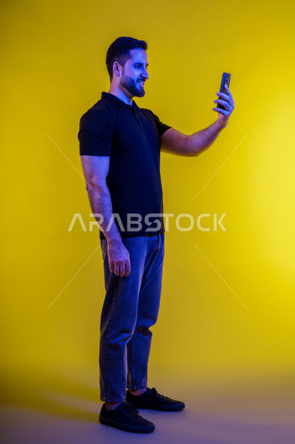 Modern digital life, interacting with technology, making a video call with friends with happy expressions, side portrait of a young Saudi Arabian man in casual wear using a mobile phone, Emirati man taking a smiling selfie, full body, yellow background