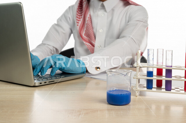 بورتريه لطالب جامعي عربي خليجي سعودي يجلس في معمل الكيمياء ،يستخدم الحاسوب المحمول لمعرفة التراكيب والتفاعلات الجزيئية ،استخدام انابيب الاختبار لاجراء التجارب في المختبر ،  التفاعلات الكيميائية ،البحث والتطوير ، مفهوم الكيمياء الحيوية