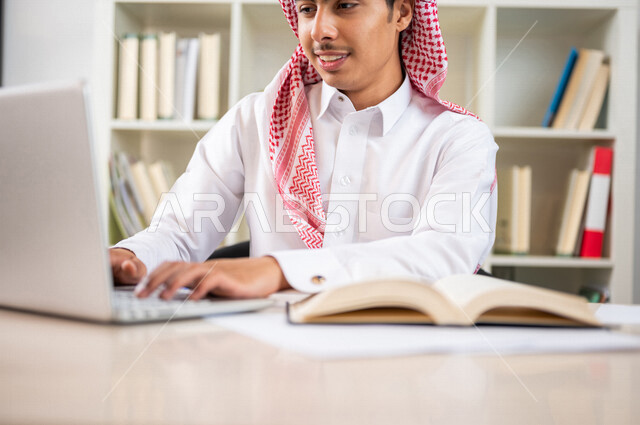 طالب جامعي عربي خليجي سعودي يرتدي الزي الخليجي التقليدي ،يقضي وقت ممتع في مكتبة الجامعة ،يستخدم الحاسوب المحمول داخل المكتبة ، استخدام التكنولوجيا في التعلم ،استعارة كتاب مناسب ، اثراء التعليم الجامعي ، البحث العلمي ، مفهوم التعليم والتعلم