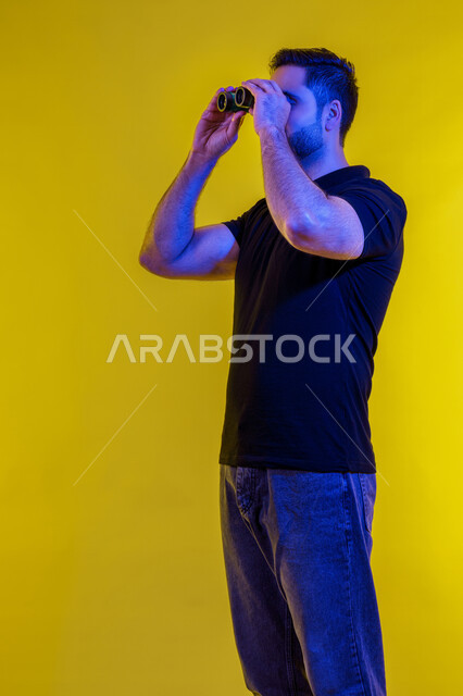 Using binoculars, love of exploration and adventure, seeing distant things using a telescope, planning for future goals and ambitions, full-length portrait of a young Arab Gulf Emirati man wearing casual clothes, a Saudi man looking through binoculars, yellow background