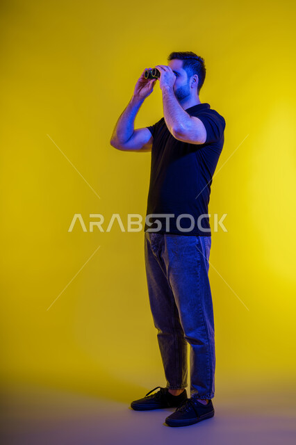 Seeing distant objects using a telescope, using binoculars and the love of exploration and adventure, planning for future goals and ambitions, full-length portrait of a young Arab Gulf Emirati man wearing casual clothes, a Saudi man looking through binoculars, yellow background