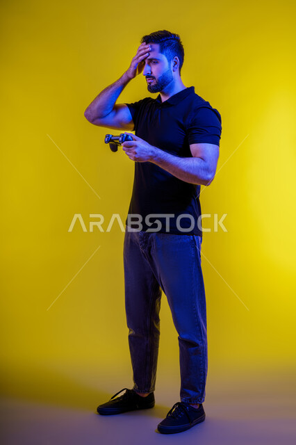 ألعاب الفيديو الترفيهية، المتعة والتسلية بالبلايستيشن، شاب عربي سعودي خليجي يمسك بيده عصا التحكم ، بورتريه لشاب اماراتي يرتدي زي كاجوال تبدو عليه ايماءات السعادة، قضاء وقت ممتع، خلفية صفراء