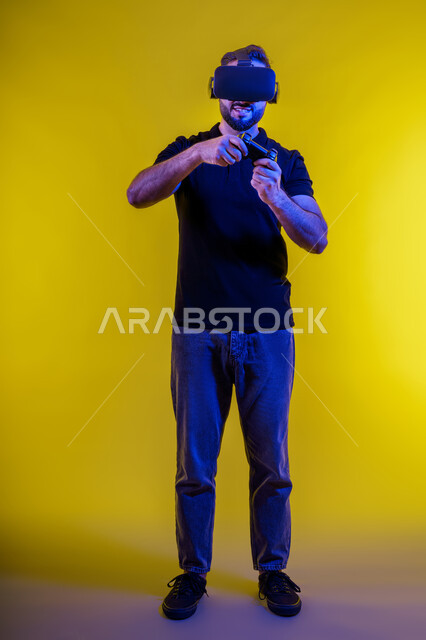 تطور التكنولوجيا بالسعودية، ألعاب البلايستيشن الترفيهية، المتعة والتسلية بألعاب الفيديو، شاب عربي سعودي خليجي يمسك بيده عصا التحكم ويرتدي نظارة الواقع الافتراضي، بورتريه لشاب اماراتي يرتدي زي كاجوال ويستمتع بتقنية الفي ار، قضاء وقت ممتع، خلفية صفراء