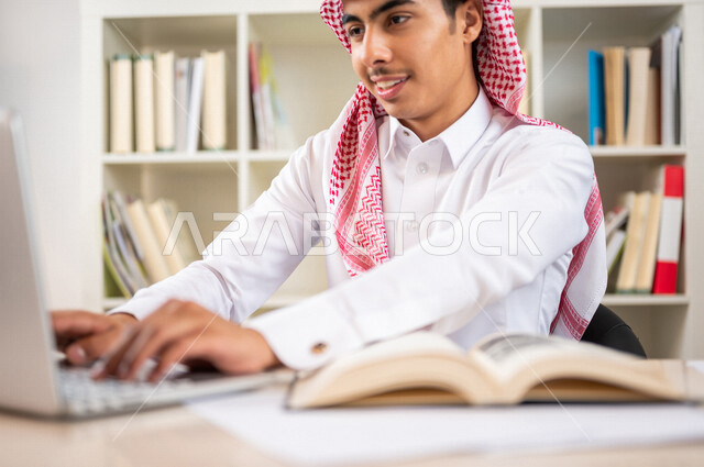 طالب جامعي عربي خليجي سعودي يرتدي الزي الخليجي التقليدي ،يقضي وقت ممتع في مكتبة الجامعة ،يستخدم الحاسوب المحمول داخل المكتبة ، استخدام التكنولوجيا في التعلم ،استعارة كتاب مناسب ، اثراء التعليم الجامعي ، البحث العلمي ، مفهوم التعليم والتعلم
