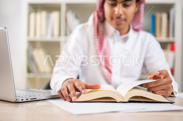 طالب جامعي عربي خليجي سعودي يرتدي الزي الخليجي التقليدي ،يقضي وقت ممتع في مكتبة الجامعة ،قراءة كتاب داخل المكتبة، تدوين الملاحظات ،استعارة كتاب مناسب ، اثراء التعليم الجامعي ، البحث العلمي ، مفهوم التعليم والتعلم