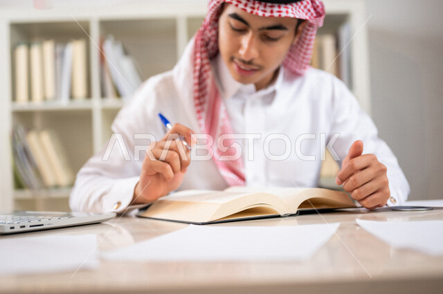 طالب جامعي عربي خليجي سعودي يرتدي الزي الخليجي التقليدي ،يقضي وقت ممتع في مكتبة الجامعة ،قراءة كتاب داخل المكتبة، تدوين الملاحظات ،استعارة كتاب مناسب ، اثراء التعليم الجامعي ، البحث العلمي ، مفهوم التعليم والتعلم