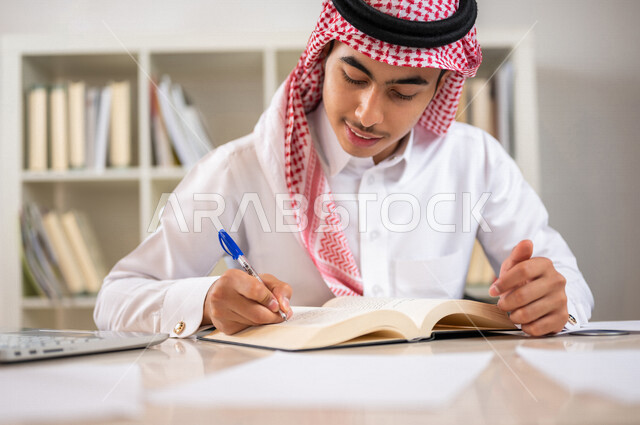 طالب جامعي عربي خليجي سعودي يرتدي الزي الخليجي التقليدي ،يقضي وقت ممتع في مكتبة الجامعة ،قراءة كتاب داخل المكتبة، تدوين الملاحظات ،استعارة كتاب مناسب ، اثراء التعليم الجامعي ، البحث العلمي ، مفهوم التعليم والتعلم