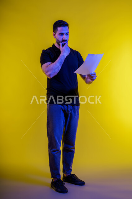 رجل عربي خليجي سعودي يرتدي الزي االكاجوال يحمل بيده كراسه تدوين الملاحظات، ايماءات وجه ويدين تدل على التفكير والحيرة، التأمل والتخطيط، افكار وحلول، التفكير بالمستقبل، بورتريه لشاب اماراتي تبدوعليه ايماءات التمعن والتركيز، خلفيه صفراء