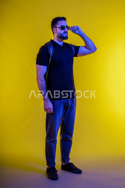 الوقوف بثقة والاهتمام بالاناقة والمظهر الخارجي العصري للرجل، شاب اماراتي خليجي عربي يرتدي الزي الكاجوال ونظارات شمسية مبتسم، بورتريه بالطول لشاب عربي سعودي تظهر عليه ايماءات الثقة والابتسام، خلفية صفراء