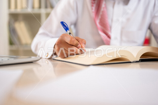 طالب جامعي عربي خليجي سعودي يرتدي الزي الخليجي التقليدي ،يقضي وقت ممتع في مكتبة الجامعة ،قراءة كتاب داخل المكتبة، تدوين الملاحظات ،استعارة كتاب مناسب ، اثراء التعليم الجامعي ، البحث العلمي ، مفهوم التعليم والتعلم