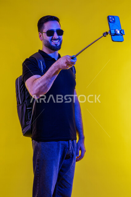 Smiling while taking selfie, contemporary techniques and interaction with technology, making a video call with friends, portrait of a young Saudi Arabian man in casual clothes, using a mobile phone, smiling Emirati man taking a selfie, yellow background