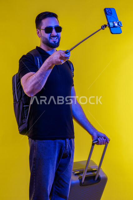 Emirati young man documenting happy moments during summer vacation, taking souvenir selfies, preparing for a fun summer trip, portrait of a smiling Saudi Arabian Gulf man in casual wear holding a suitcase using a mobile phone, yellow background