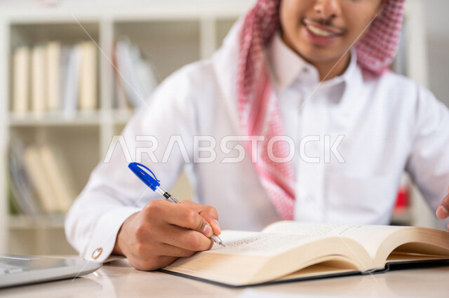 طالب جامعي عربي خليجي سعودي يرتدي الزي الخليجي التقليدي ،يقضي وقت ممتع في مكتبة الجامعة ،قراءة كتاب داخل المكتبة، تدوين الملاحظات ،استعارة كتاب مناسب ، اثراء التعليم الجامعي ، البحث العلمي ، مفهوم التعليم والتعلم