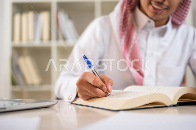 طالب جامعي عربي خليجي سعودي يرتدي الزي الخليجي التقليدي ،يقضي وقت ممتع في مكتبة الجامعة ،قراءة كتاب داخل المكتبة، تدوين الملاحظات ،استعارة كتاب مناسب ، اثراء التعليم الجامعي ، البحث العلمي ، مفهوم التعليم والتعلم