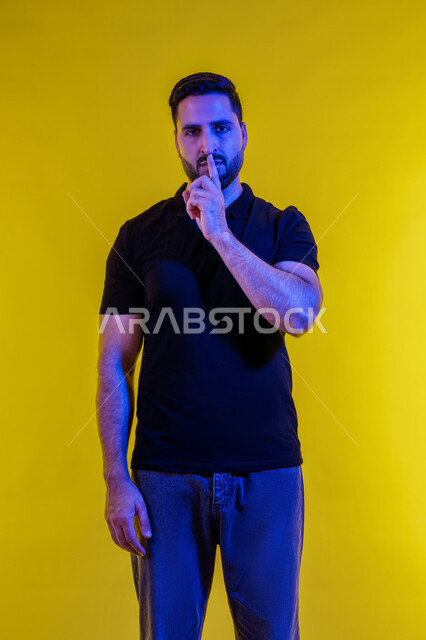 Stop talking and lower your voice, calm and quiet and do not disturb, portrait of a young Saudi Arabian Gulf man in casual clothes standing straight and putting his index finger on his mouth, Emirati man asking for silence and quiet, full body image, yellow background