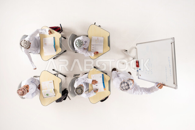 Interest in the educational process, explanation on the whiteboard, group activities for teaching students in Omani schools, Gulf Arab Omani students wearing traditional dishdasha and kummah sitting on school benches, vertical overhead portrait of a Gulf Arab Omani teacher giving lessons, white background
