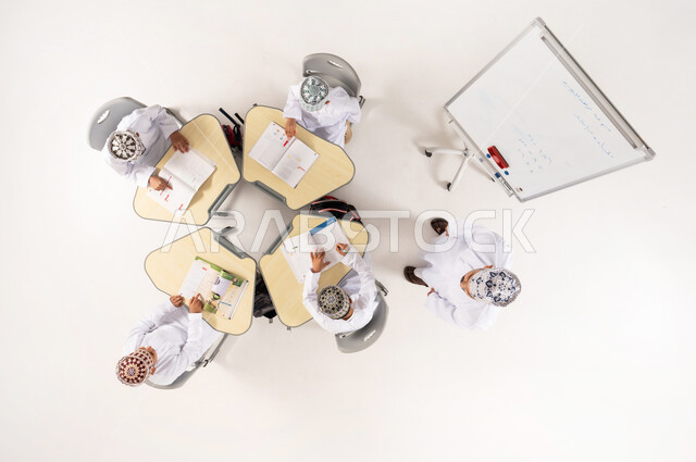 Interest in the educational process, explanation on the whiteboard, group activities for teaching students in Omani schools, Gulf Arab Omani students wearing traditional dishdasha and kummah sitting on school benches, vertical overhead portrait of a Gulf Arab Omani teacher giving lessons, white background