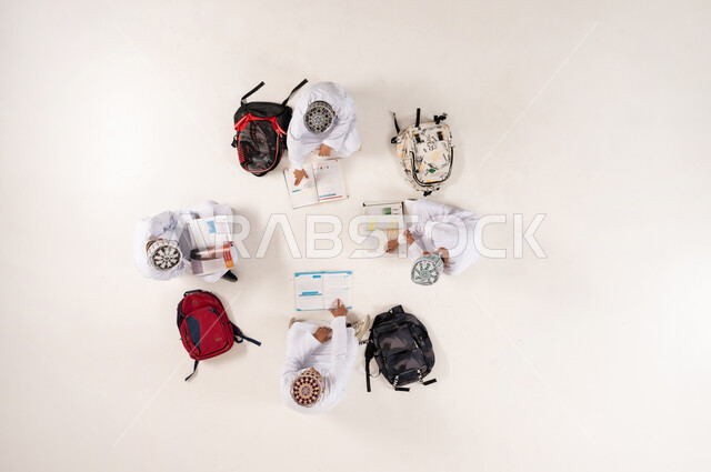 Expressions of concentration and engagement in solving assignments, developing creative skills, vertical overhead portrait of a group of Omani Gulf Arab boys wearing traditional dishdashas and kummah sitting on the floor, enjoying group recreational activities, writing homework with friends, white background