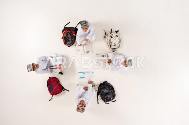 Writing homework with friends, expressions of concentration and engagement in solving homework, developing creative skills, vertical overhead portrait of a group of Omani Gulf Arab boys wearing traditional dishdasha and kummah sitting on the floor, enjoying group recreational activities, white background