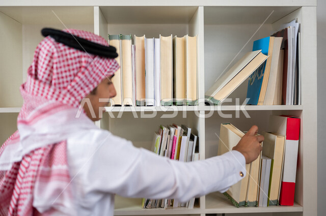 طالب جامعي عربي خليجي سعودي يرتدي الزي الخليجي التقليدي ،يقضي وقت ممتع في مكتبة الجامعة ،البحث بين الكتب، استعارة كتاب مناسب ، اثراء التعليم الجامعي ، البحث العلمي ، مفهوم التعليم والتعلم