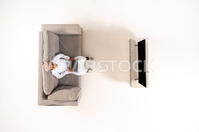 Entertaining video games, vertical overhead portrait of an Arab Gulf Omani boy wearing a traditional dishdasha and kummah sitting on a sofa in the living room working on a laptop, blank black screen on TV, entertainment using modern technology, white background