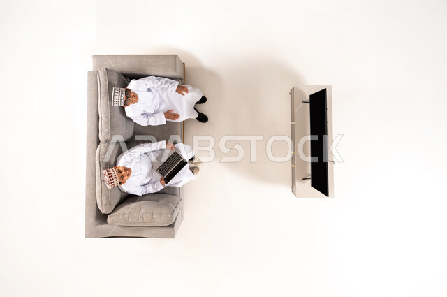 Watching TV shows, practicing fun hobbies, using modern technology during breaks, entertainment and fun during free time via laptop, vertical overhead portrait of an Omani Gulf Arab family wearing a dishdasha and kummah sitting in the living room, white background