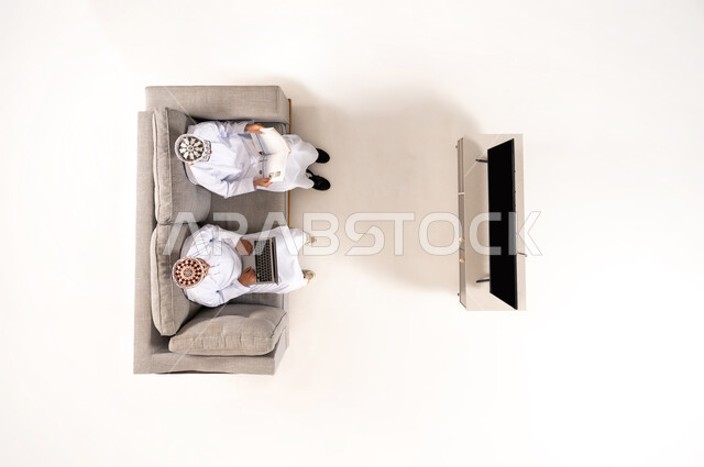 Watching TV shows, practicing fun hobbies, using modern technology during breaks, entertainment and fun during free time via laptop, vertical overhead portrait of an Omani Gulf Arab family wearing a dishdasha and kummah sitting in the living room, white background