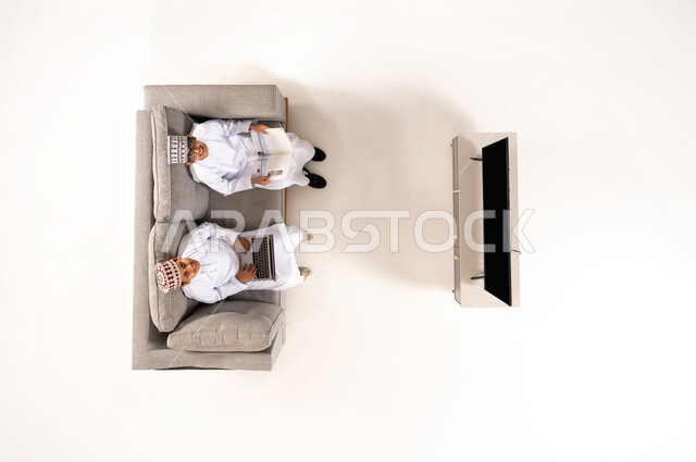 Practicing entertaining hobbies, using modern technology during break time, entertainment and fun during free time via laptop, vertical overhead portrait of an Omani Gulf Arab family wearing a dishdasha and a kummah sitting in the living room, watching TV programs, white background