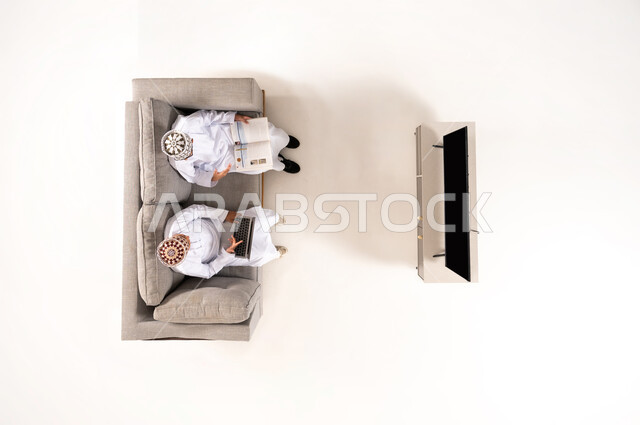 Watching TV shows, practicing fun hobbies, using modern technology during breaks, entertainment and fun during free time via laptop, vertical overhead portrait of an Omani Gulf Arab family wearing a dishdasha and kummah sitting in the living room, white background