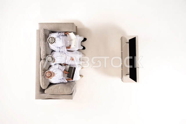 Watching TV shows, practicing fun hobbies, using modern technology during breaks, entertainment and fun during free time via laptop, vertical overhead portrait of an Omani Gulf Arab family wearing a dishdasha and kummah sitting in the living room, white background