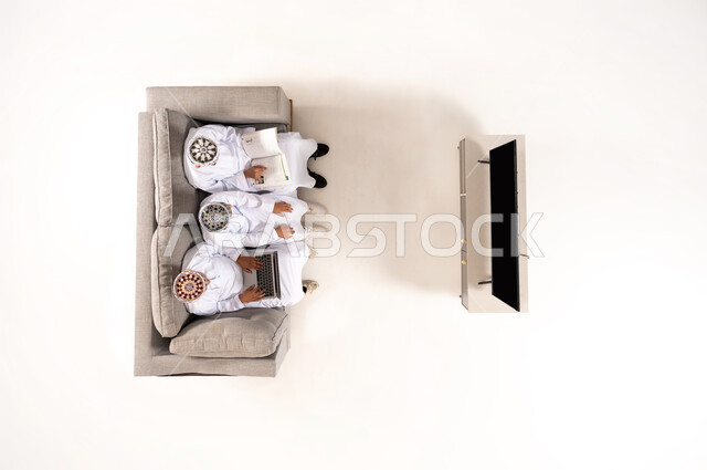 Practicing entertaining hobbies, using modern technology during break time, entertainment and fun during free time via laptop, vertical overhead portrait of an Omani Gulf Arab family wearing a dishdasha and a kummah sitting in the living room, watching TV programs, white background