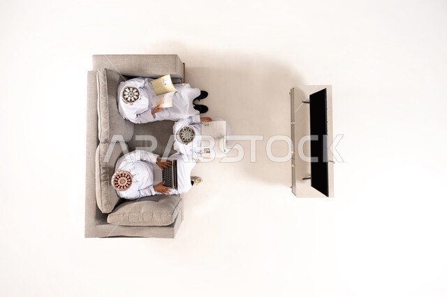 Watching TV shows, practicing fun hobbies, using modern technology during breaks, entertainment and fun during free time via laptop, vertical overhead portrait of an Omani Gulf Arab family wearing a dishdasha and kummah sitting in the living room, white background