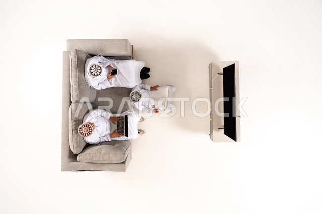 Watching TV shows, practicing fun hobbies, using modern technology during breaks, entertainment and fun during free time via laptop, vertical overhead portrait of an Omani Gulf Arab family wearing a dishdasha and kummah sitting in the living room, white background