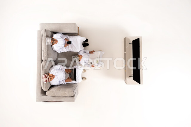 Practicing entertaining hobbies, using modern technology during break time, entertainment and fun during free time via laptop, vertical overhead portrait of an Omani Gulf Arab family wearing a dishdasha and a kummah sitting in the living room, watching TV programs, white background