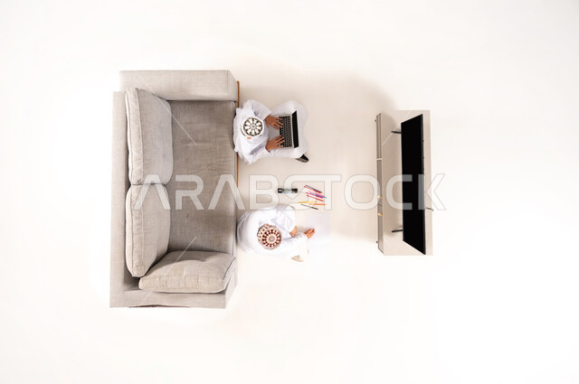 Practicing entertaining hobbies, using modern technology during break time, entertainment and fun during free time via laptop, vertical overhead portrait of an Omani Gulf Arab family wearing a dishdasha and a kummah sitting in the living room, watching TV programs, white background