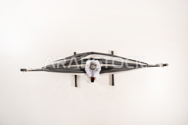 دمج التقنيات الحديثة بالتسلية والمرح، قضاء أوقات ممتعة في العطلة، مفهوم الاسترخاء والاستجمام، بورتريه علوي عامودي لولد عربي خليجي عماني يرتدي الدشداشة والكمة التقليدية يجلس على أرجوحة قماشية معلقة يقوم باستخدام الهاتف المحمول، خلفية بيضاء