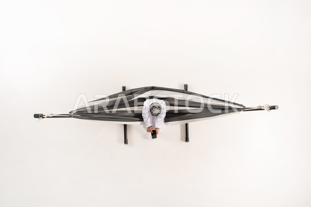 Combining modern technology with entertainment and fun, having a good time on vacation, the concept of relaxation and recreation, a vertical overhead portrait of an Arab Gulf Omani boy wearing a traditional dishdasha and kummah sitting on a hanging cloth swing using a mobile phone, white background