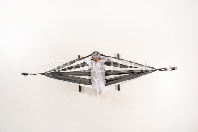The need for rest and tranquility, the concept of relaxation and recreation, a vertical overhead portrait of an Omani Gulf Arab boy wearing the traditional dishdasha and kummah sitting on a cloth swing, white background