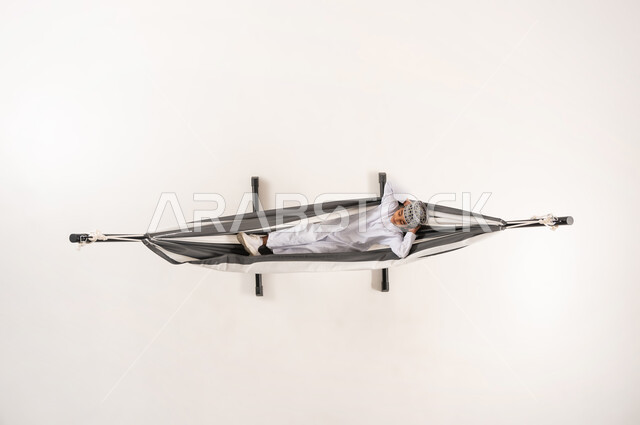 The need for rest and tranquility, the concept of relaxation and recreation, a vertical overhead portrait of an Omani Gulf Arab boy wearing the traditional dishdasha and kummah sitting on a cloth swing, white background