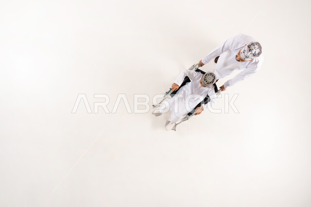 الدعم والتشجيع العائلي لأصحاب الهمم, تحسين حياة ذوي الإعاقة الحركية على التنقل, بورتريه علوي عامودي لرجل عربي خليجي عماني يرتدي الدشداشة والكمة التقليدية يقوم بمساعدة ابنه من ذوي الاحتياجات الخاصة، استخدام الكرسي المتحرك، خلفية بيضاء
