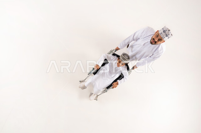 الدعم والتشجيع العائلي لأصحاب الهمم, تحسين حياة ذوي الإعاقة الحركية على التنقل, بورتريه علوي عامودي لرجل عربي خليجي عماني يرتدي الدشداشة والكمة التقليدية يقوم بمساعدة ابنه من ذوي الاحتياجات الخاصة، استخدام الكرسي المتحرك، خلفية بيضاء