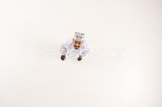 Concern for outward appearance, concept of masculinity and elegance, standing upright and upright, vertical overhead portrait of a smiling Omani Gulf Arab man wearing the traditional dishdasha and kummah, looking at the camera with expressions and gestures of pleasure and happiness, white background